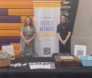 Two women standing behind a table smiling in front of a World Affairs Council sign.